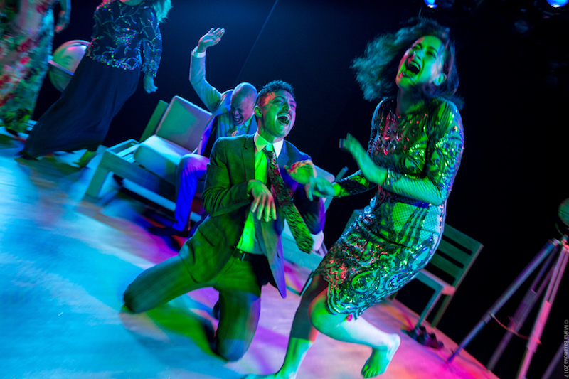 The performers in mid cackle. One, in a plaid suit sits on his knees and tilts his head back, another sits on the couch about to slap his knee and women with her mouth agape